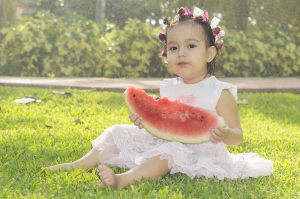 Menina branca e bonita, com uma faixa de flores na cabeça, sentada no relvado verde de um jardim, a comer uma fatia de melancia que ela segura nas mãos. — Fotografia de Stock