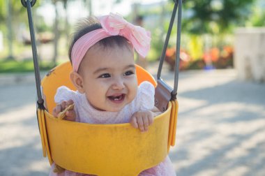 Bir yaşında bir bebek, sarı salıncak ve pembe fiyonk, elinde bir kurabiye, mutlu bir şekilde gülüyor ve iki küçük diş çıkıyor, odaklanmamış arka plan.