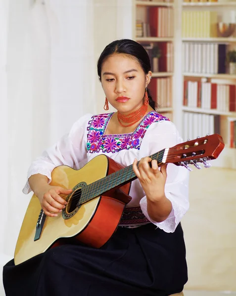 stock image Portrait of young pretty woman wearing beautiful traditional andean clothing, sitting down with acoustic guitar playing, bookshelves background