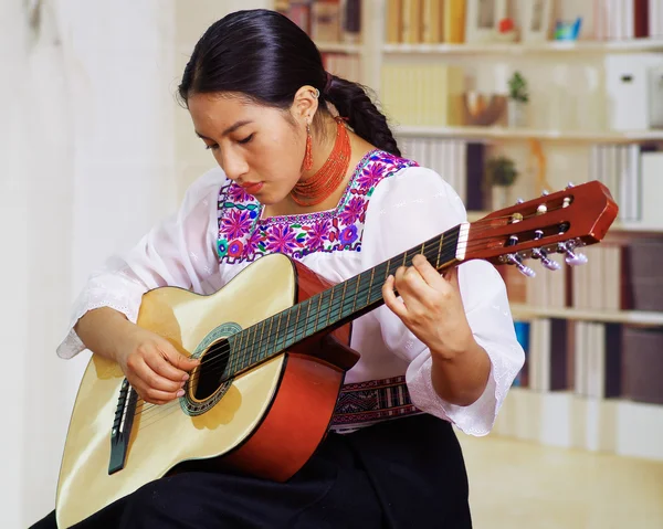 Portret van jonge mooie vrouw het dragen van prachtige traditionele Andes kleding, zittend met akoestische gitaar spelen, bookplanken achtergrond — Stockfoto