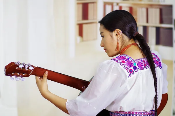 Портрет молодой красивой женщины в красивой традиционной андской одежде, сидящей за игрой на акустической гитаре, на книжных полках — стоковое фото