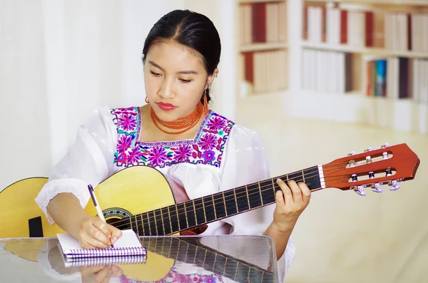 Portrait of young pretty woman wearing beautiful traditional andean clothing, sitting down with acoustic guitar playing, bookshelves background — Stock Photo, Image