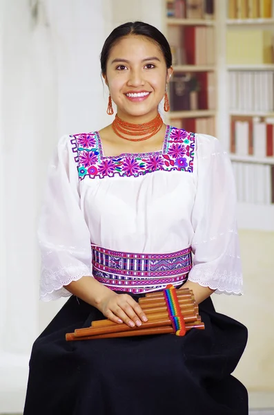 Porträtt av ung vacker kvinna som bär vackra traditionella andinska kläder, sitta med och spela munspel, bokhyllor bakgrund — Stockfoto