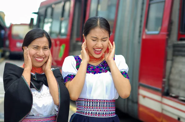Hermosa madre e hija hispanas vistiendo ropa andina tradicional, de pie en la plataforma de la estación cubriendo las orejas mientras pasa el autobús rojo, ambiente al aire libre —  Fotos de Stock