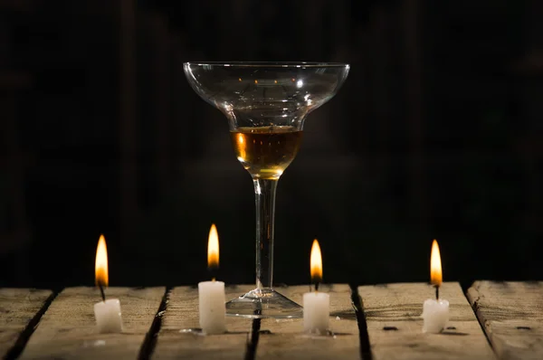Quattro candele di cera bianca sedute su una superficie di legno che brucia, bicchiere da cocktail posto dietro, sfondo nero — Foto Stock