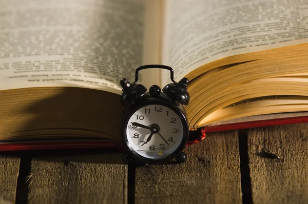 Thick livro deitado aberto na superfície de madeira, à moda antiga relógio de mesa de noite sentado ao lado dele, tiro conceito mágico — Fotografia de Stock