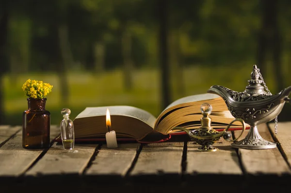 Thick book lying open on wooden surface, small brown bottle with flowers, Aladin style lamps and wax candle next to it, magic concept shoot — Stock Photo, Image