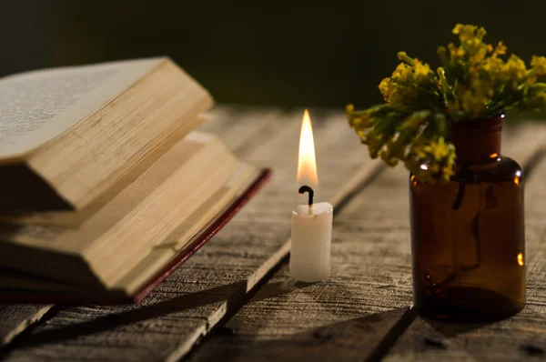 Libro grueso acostado abierto en la superficie de madera, vela de cera y una pequeña botella con flores amarillas sentadas a su lado, hermoso escenario de luz nocturna, sesión de concepto mágico — Foto de Stock