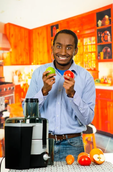 Homem encantador vestindo jeans jeans combinando camisa, segurando maçã e tomate, posando feliz para câmera, fabricante de suco profissional colocado na mesa na frente, conceito de estilo de vida saudável — Fotografia de Stock