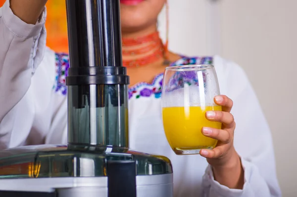 Primeros planos de las manos de la mujer sosteniendo vidrio con jugo de naranja, otra mano descansando en la máquina, concepto de estilo de vida saludable — Foto de Stock