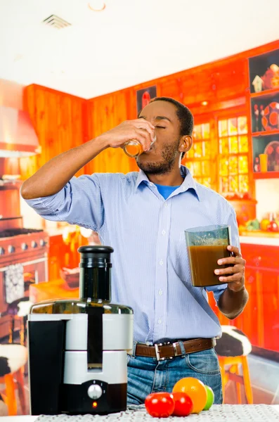 Hombre vistiendo vaqueros camisa a juego de pie dentro de la cocina junto a la máquina de jugo, beber de vidrio, concepto de estilo de vida saludable —  Fotos de Stock