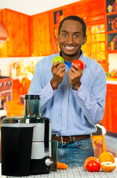 Чарівний чоловік у джинсовій сорочці, що відповідає джинсам, тримає яблуко та помідор, щасливо позує на камеру, професійний виробник соків, розміщений на столі спереду, концепція здорового способу життя — стокове фото