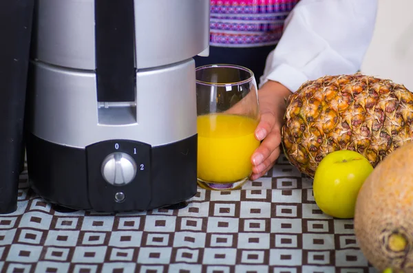 Closeup fabricante de suco parcialmente visível sentado na mesa ao lado de abacaxi, pêra e maçã, mão segurando em vidro de líquido amarelo — Fotografia de Stock