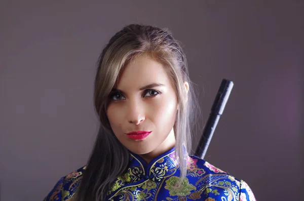 Samurai vrouw gekleed in traditionele kleurrijke bloem patroon Aziatische zijden jurk, houdt van arm over de schouder grijpen zwaard verborgen achter de rug, geconfronteerd met camera, ninja concept — Stockfoto