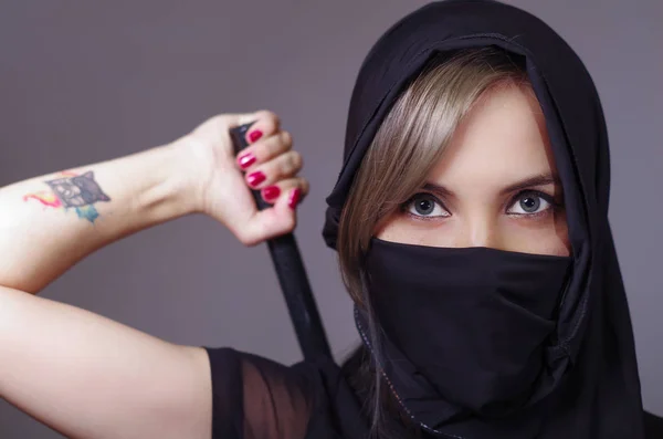 Mujer samurai vestida de negro con velo a juego cubriendo la cara, sosteniendo el brazo en la espada oculta detrás de la espalda, frente a la cámara, concepto ninja — Foto de Stock