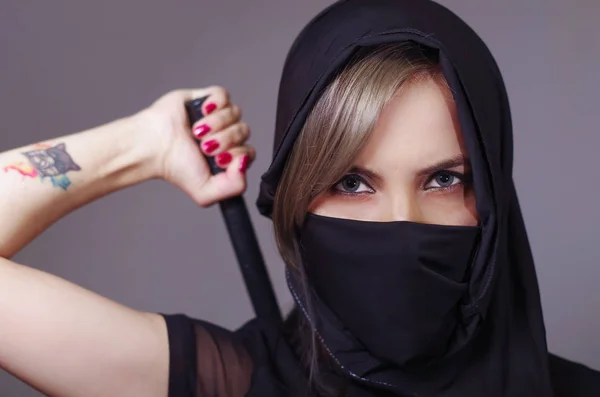 Mujer samurai vestida de negro con velo a juego cubriendo la cara, sosteniendo el brazo en la espada oculta detrás de la espalda, frente a la cámara, concepto ninja — Foto de Stock