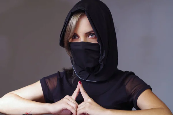 Mujer samurai en la cabeza vestida de negro con velo a juego cubriendo la cara, apoyando los brazos en el escritorio y tocando las yemas de los dedos uno contra el otro, frente a la cámara, concepto ninja — Foto de Stock