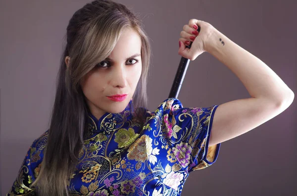 Samurai vrouw gekleed in traditionele kleurrijke bloem patroon Aziatische zijden jurk, houdt van arm over de schouder grijpen zwaard verborgen achter de rug, geconfronteerd met camera, ninja concept — Stockfoto