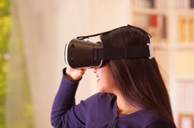 Young brunette woman wearing virtual reality goggles experiencing future technology, interacting and smiling while playing, domestic background,vr concept clipart