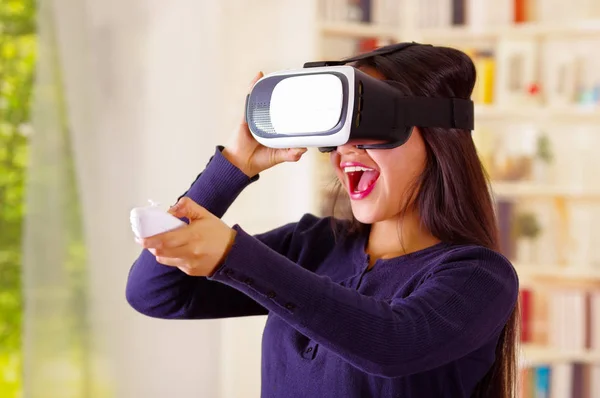 Mujer morena joven con gafas de realidad virtual experimentando la tecnología del futuro, interactuando y sonriendo mientras juega, fondo doméstico, concepto vr —  Fotos de Stock