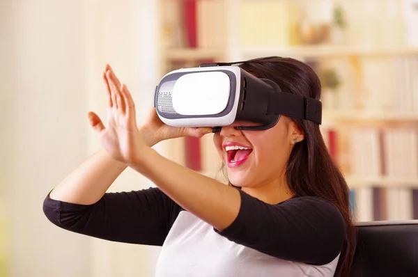 Mujer morena joven con gafas de realidad virtual experimentando la tecnología del futuro, interactuando y sonriendo mientras juega, fondo doméstico, concepto vr —  Fotos de Stock