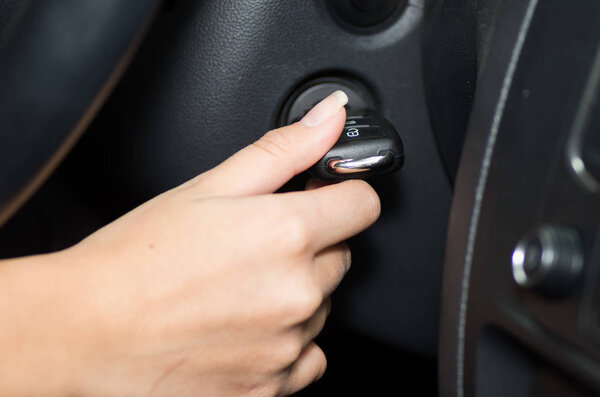 Closeup inside vehicle of hand holding key in ignition, steering wheel and black interior background, female driver concept