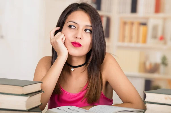 年轻的黑发女人，穿着粉红色顶坐桌边有一摞书放在上面，下巴上的手，沉思的面部表情，学生概念 — 图库照片