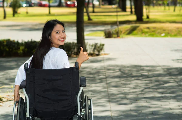 Молодая брюнетка женщина сидит в инвалидной коляске улыбаясь с позитивным настроем, на открытом воздухе, концепция физического восстановления — стоковое фото