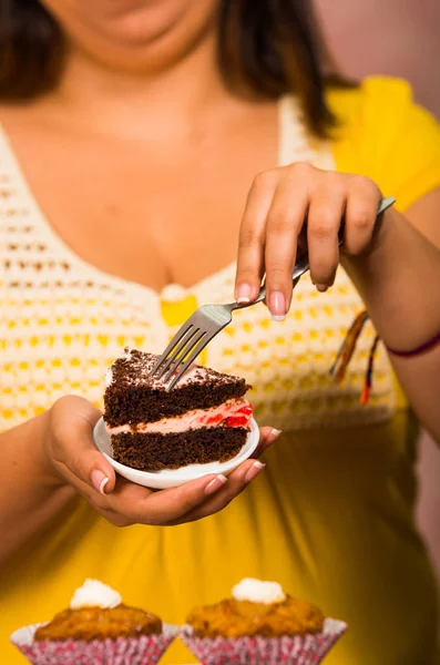 Woman holding small plate of chocolate cake with cream filling, grabbing bite using fork, showing to camera, pastry concept — Stock Photo, Image
