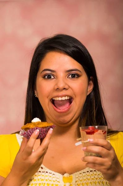 Donna bruna che tiene in mano delizioso muffin marrone con crema di condimento, vetro di mousse in altra mano, grande sorriso e pronto a prendere un morso, concetto di pasticceria — Foto Stock