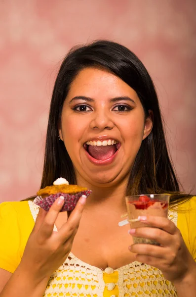Brunette woman holding delicious brown colored muffin with cream topping, glass of mousse in other hand, big smile and ready to take a bite, pastry concept — Stock Photo, Image
