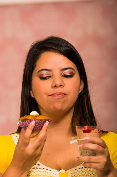 Brunette woman holding delicious brown colored muffin with cream topping, glass of mousse in other hand, big smile and ready to take a bite, pastry concept — Stock Photo, Image