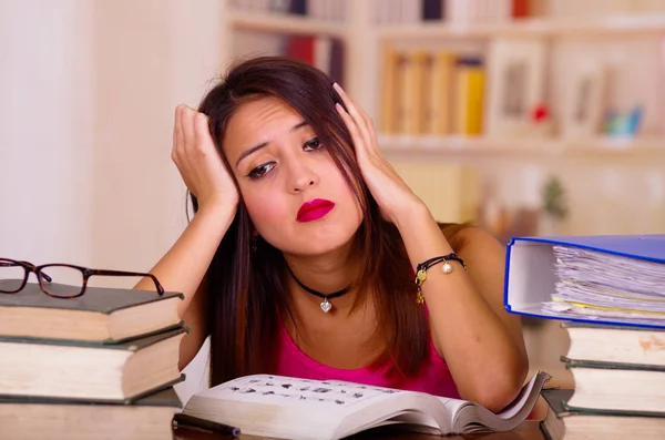 Giovane donna bruna vestita con top rosa seduta alla scrivania con una pila di libri appoggiati su di essa, appoggiata a testa in mano, espressione facciale stanca, concetto di studente — Foto Stock