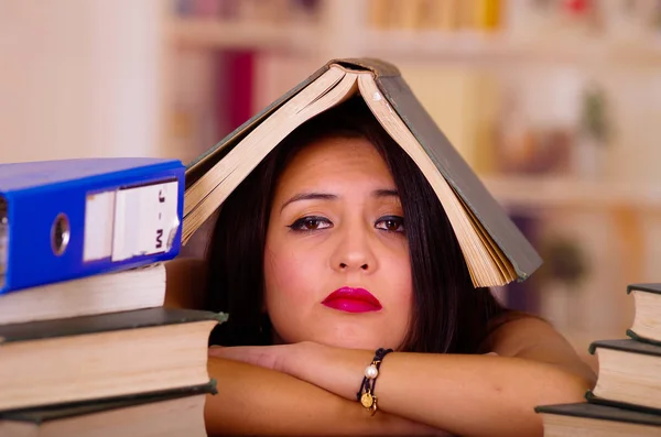 Ung brunett kvinna som bär rosa topp ligga böjd över skrivbordet med trave böcker, öppna boken placeras på huvudet, trött ansiktsuttryck och kroppsspråk, student koncept — Stockfoto
