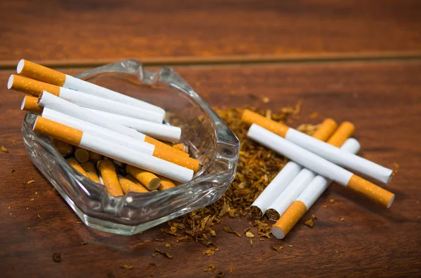 Cigarettes and tobacco lying inside and around glass ash tray on wooden surface, seen from above, anti smoking concept — Stock Photo, Image