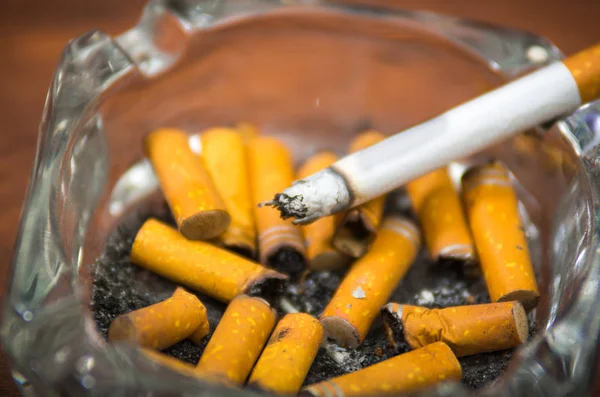 Cigarettes and tobacco lying inside and around glass ash tray on wooden surface, seen from above, anti smoking concept — Stock Photo, Image