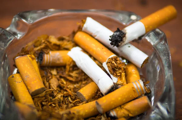 Cigarrillos y tabaco que se encuentran dentro y alrededor del cenicero de vidrio en la superficie de madera, visto desde arriba, concepto antitabaco — Foto de Stock