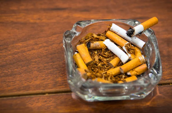 Cigarettes and tobacco lying inside and around glass ash tray on wooden surface, seen from above, anti smoking concept — Stock Photo, Image