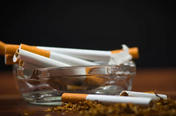 Nahaufnahme Zigaretten und Tabak in und um Glasaschenbecher auf Holzoberfläche liegend, Anti-Raucher-Konzept — Stockfoto