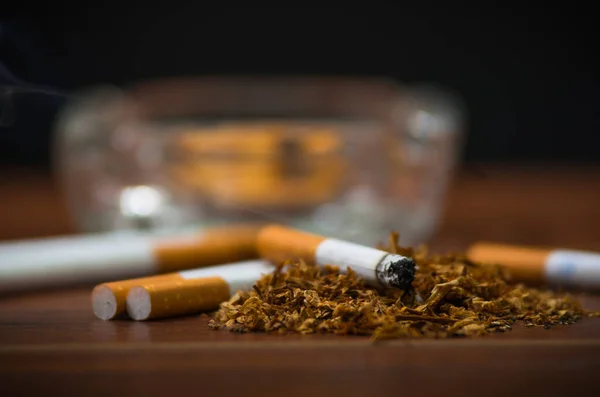 Closeup cigarettes and tobacco lying inside and around glass ash tray on wooden surface, anti smoking concept — Stock Photo, Image