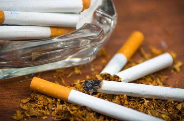 Closeup cigarettes and tobacco lying inside and around glass ash tray on wooden surface, anti smoking concept — Stock Photo, Image