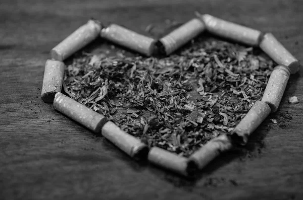Cigarette butts shaped into a heart lying on wooden surface, tobacco spread around middle, seen from above, black and white edition — Stock Photo, Image