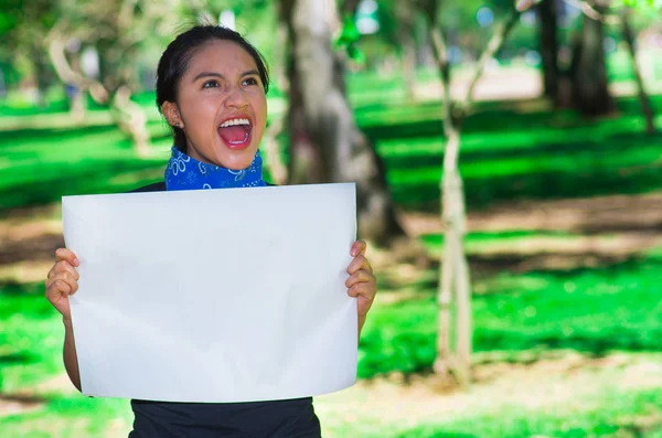 Mujer morena joven vistiendo pañuelo azul alrededor del cuello, sosteniendo afiche blanco cantando al aire libre frente a la cámara, concepto de protesta activista —  Fotos de Stock