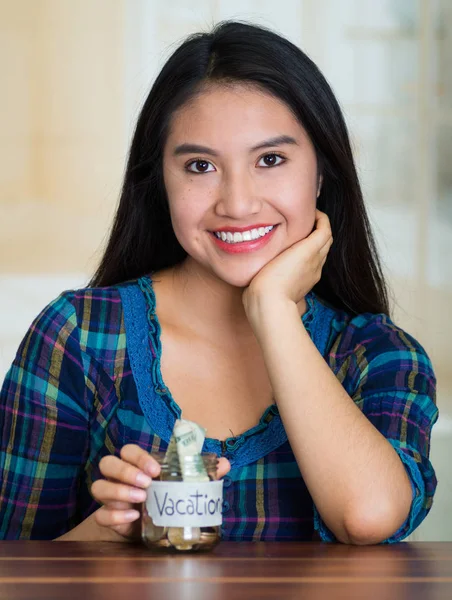 Giovane donna bruna seduta di fronte alla macchina fotografica, con dentro un barattolo di vetro con monete, etichetta che legge vacanze, sorridente felicemente — Foto Stock