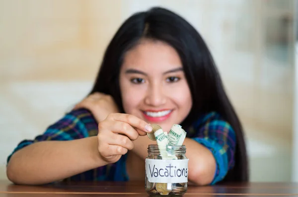 Junge brünette Frau setzt sich vor die Kamera, steckt Münzen in Glasgefäße mit Geld darin, beschriftet Urlaube, lächelt glücklich — Stockfoto