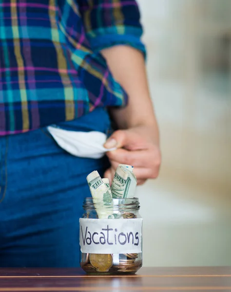 Frau steht vor Kamera und hält leere Jeanstasche hoch, Glasgefäß mit Geld sitzt auf Schreibtisch davor, Etikett liest Urlaub — Stockfoto