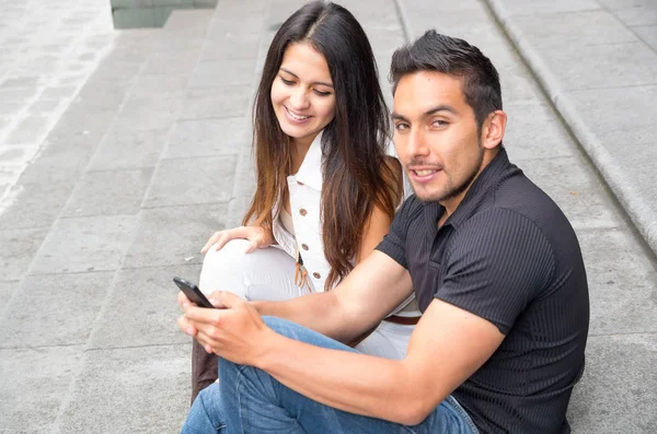 Encantadora pareja joven sentada en la construcción de escalones utilizando el teléfono móvil e interactuando felizmente, concepto turístico urbano —  Fotos de Stock