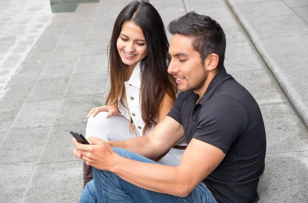 Charming young couple sitting on building steps using mobile phone and interacting happily, urban tourist concept — Stock Photo, Image