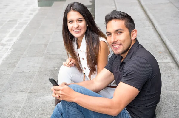 Encantadora pareja joven sentada en la construcción de escalones utilizando el teléfono móvil e interactuando felizmente, concepto turístico urbano —  Fotos de Stock