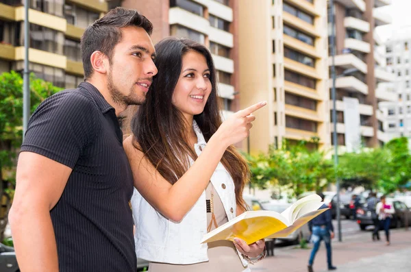 Encantadora pareja joven parada fuera en el entorno urbano, sosteniendo libro abierto y señalando con el dedo, concepto turístico urbano — Foto de Stock